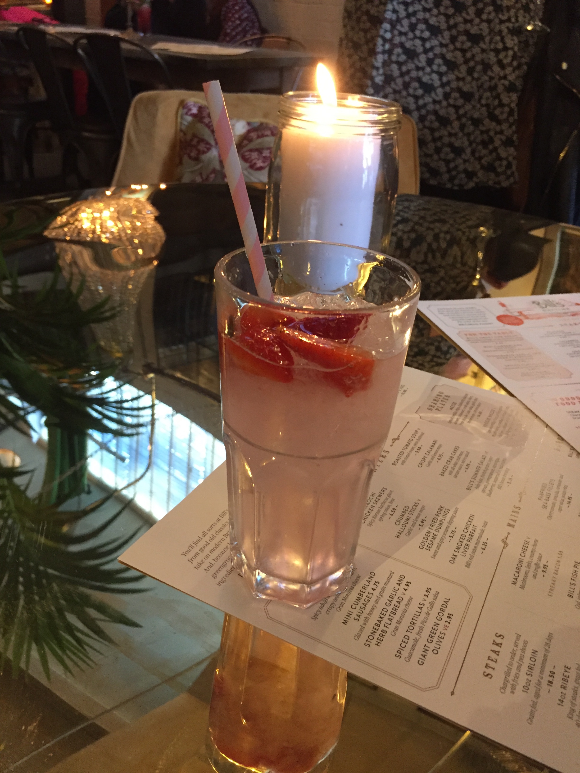 a glass of pink drink with straw and ice on a table with a candle